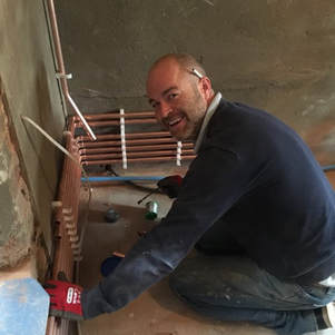 Arco Director working across Essex. Arco member, Craig is sat on the floor of a room touching copper piping that's attached to the wall. He is turning and smiling towards the camera.